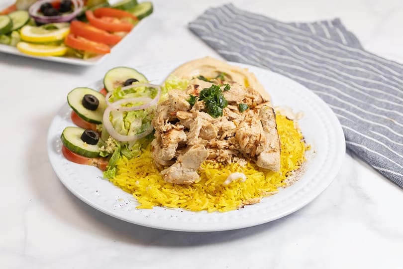 Chicken shawarma plate with yellow rice, hummus, and a side salad with cucumbers, tomatoes, and olives.
