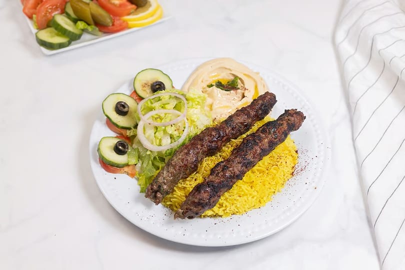 Two grilled meat skewers on yellow rice with hummus, salad, and a side of pickles and tomatoes.