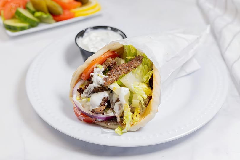 Gyro sandwich with meat, lettuce, tomatoes, onions, and white sauce, with a side of tzatziki.